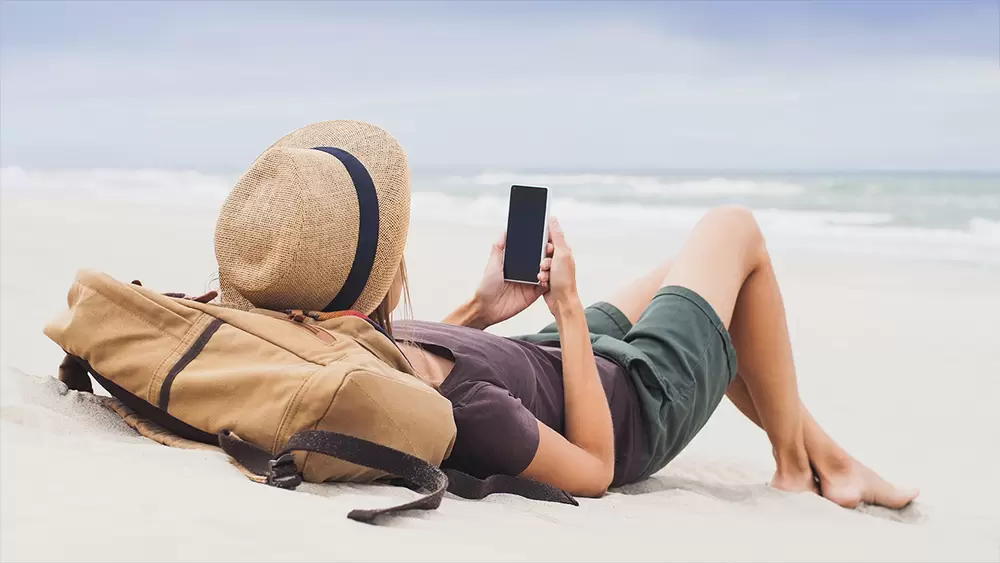 Semesterklädd kvinna ligger på stranden och tittar på sin mobiltelefon